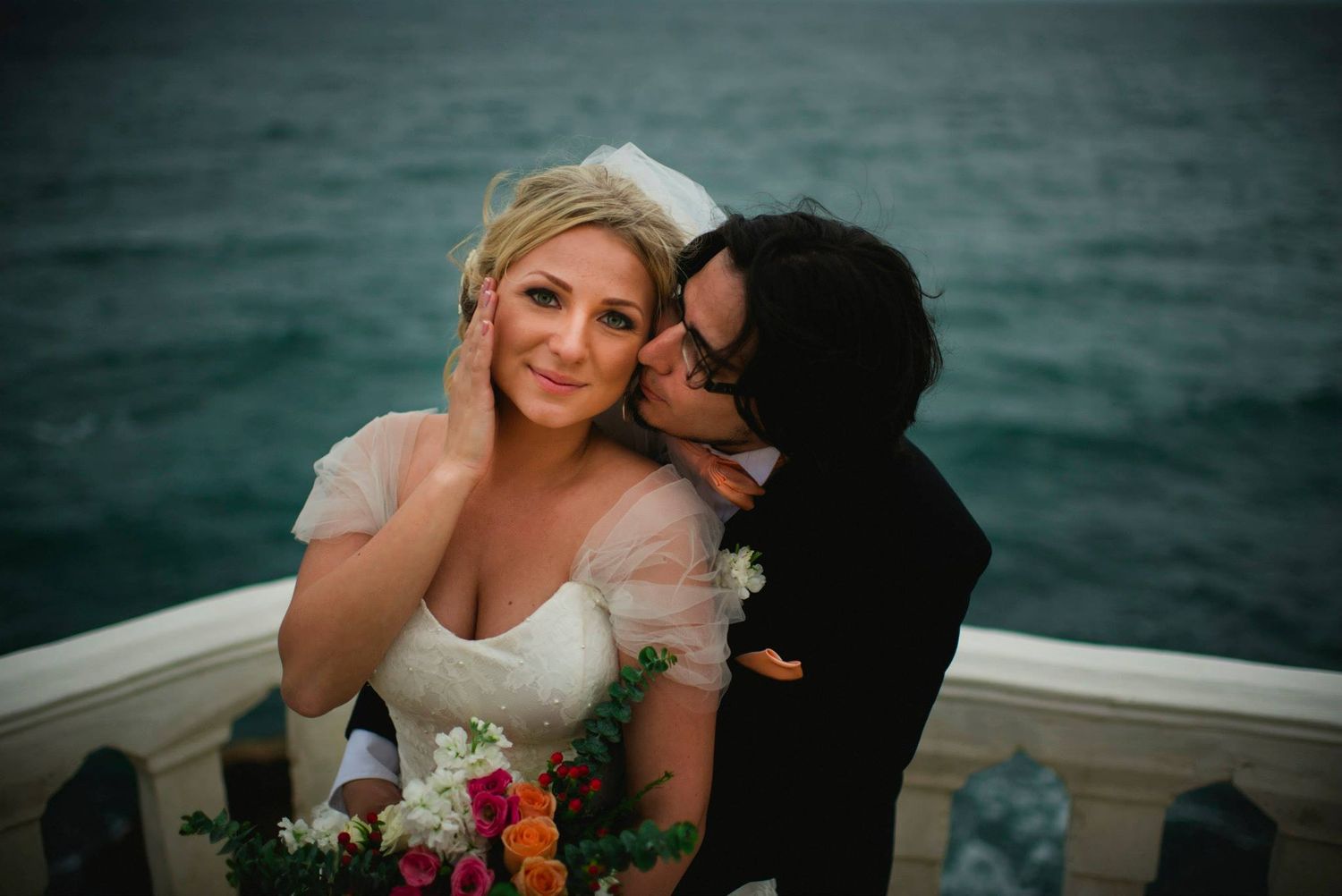 Xenia Helix holding a colorful bouquet and receiving a kiss on the cheek from the groom on the balcony over the ocean.