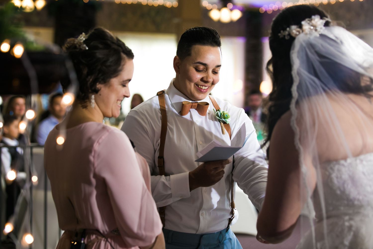 Bride in suspenders and bow tie reads vows at an indoor wedding ceremony with the other bride, at Monoosnock Country Club, Leominster, MA