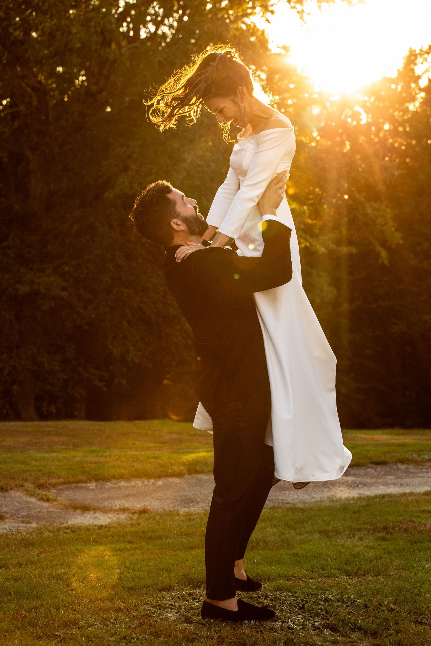 Groom lifts bride in white dress at golden sunset in lush green park during wedding celebration, Xenia Helix Photography, Boston, MA