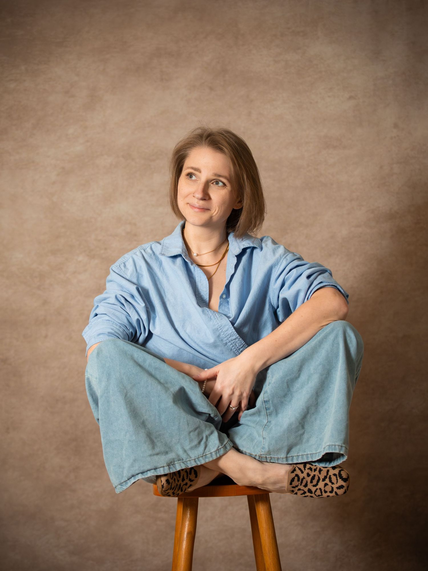 Xenia Helix, Boston wedding photographer, in blue denim shirt and wide-leg jeans sits cross-legged on a wooden stool, neutral backdrop