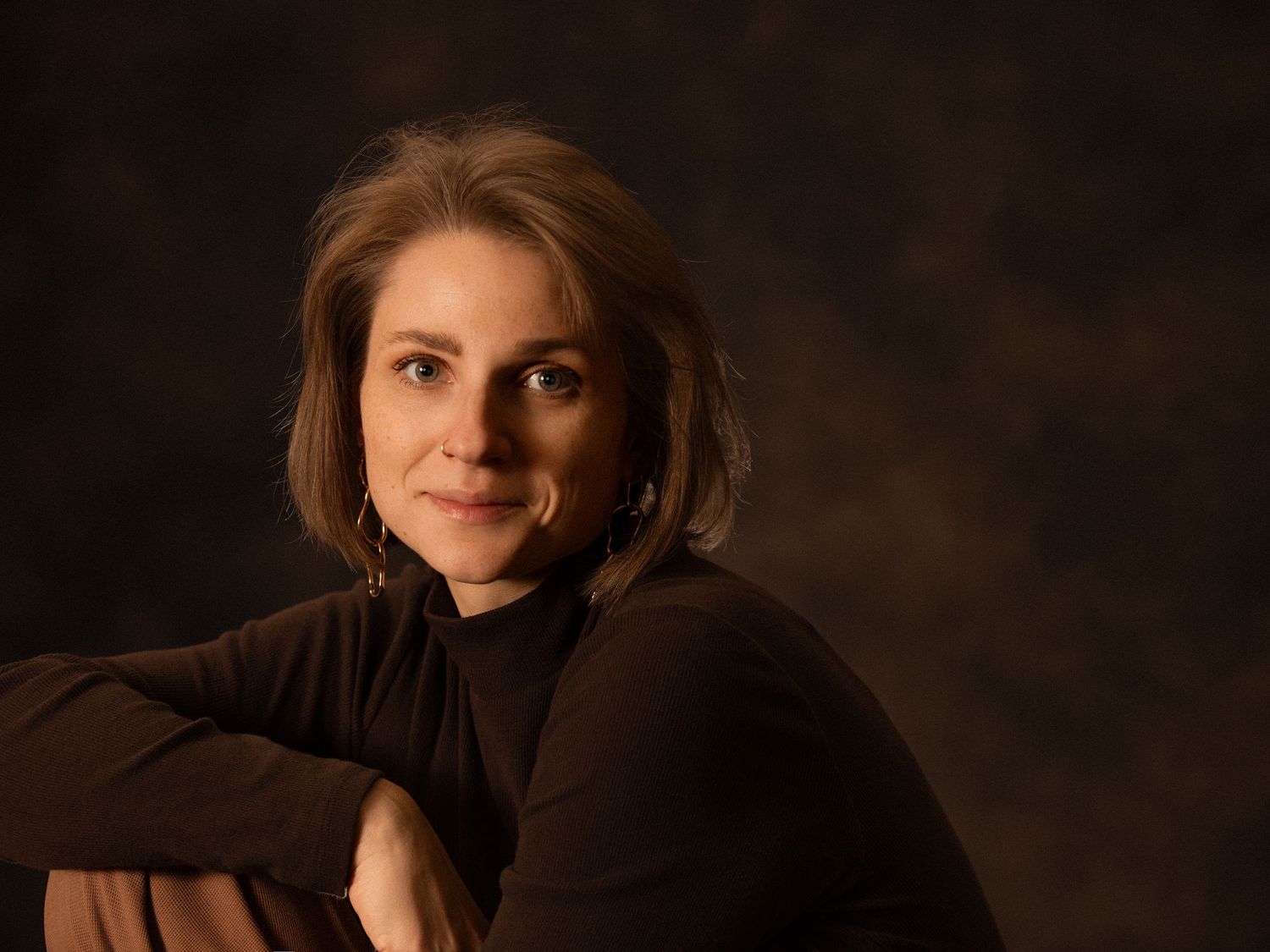 Xenia Helix, Boston wedding photographer in a brown turtleneck, posing with arms crossed on a dark background.