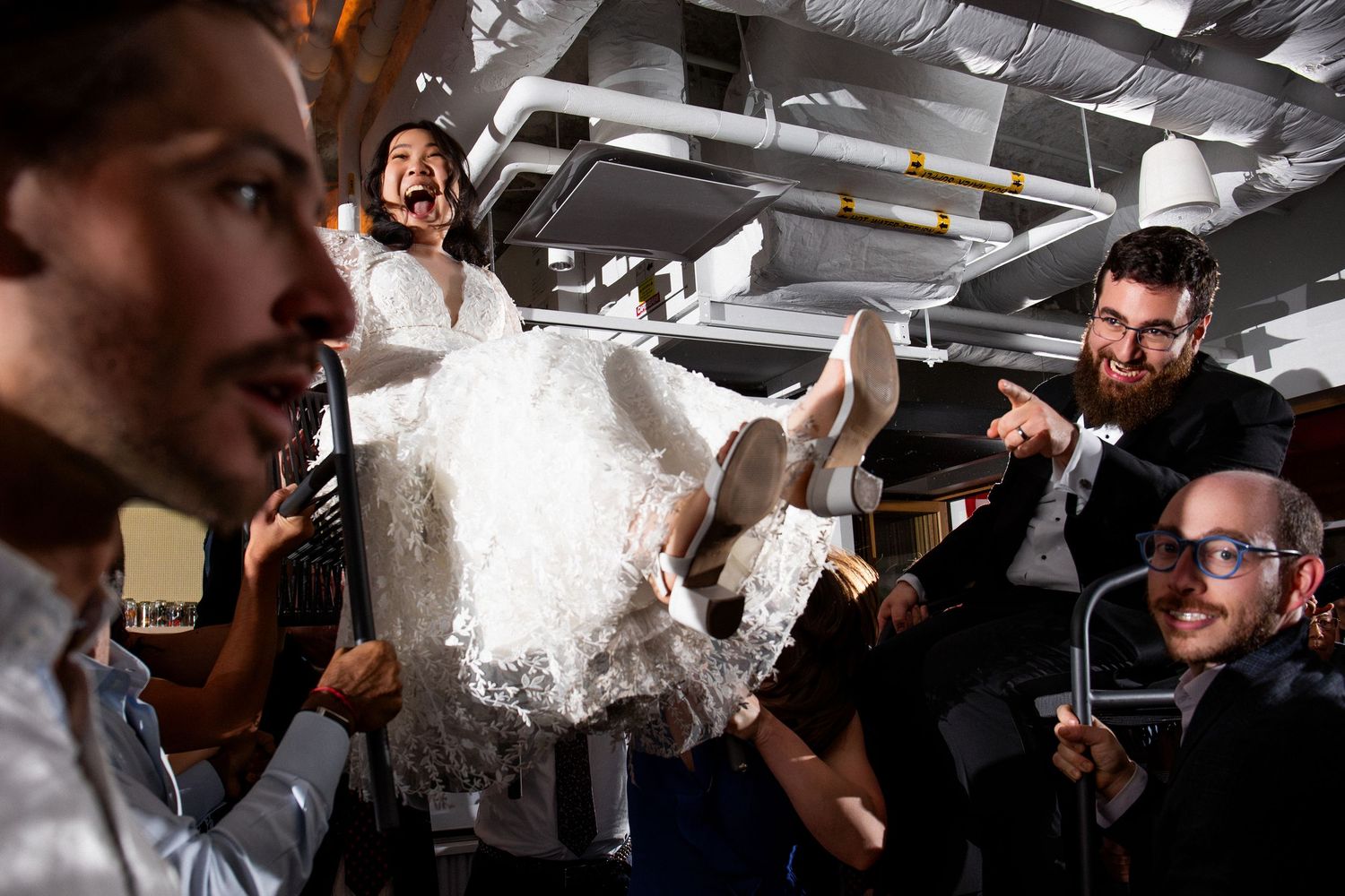 Laughing bride in white dress hoisted up by smiling groomsmen during energetic wedding hora, Mothership Alewife, Cambridge, MA