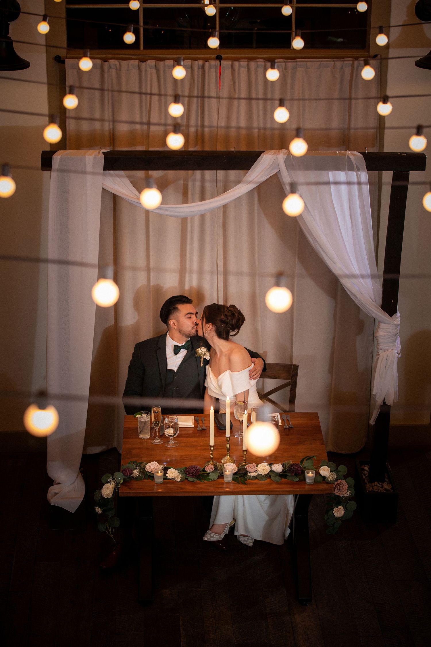 Bride and groom kiss at sweetheart table surrounded by warm Edison bulb string lights, top view, Grove at Briar Barn Inn, Rowley, MA