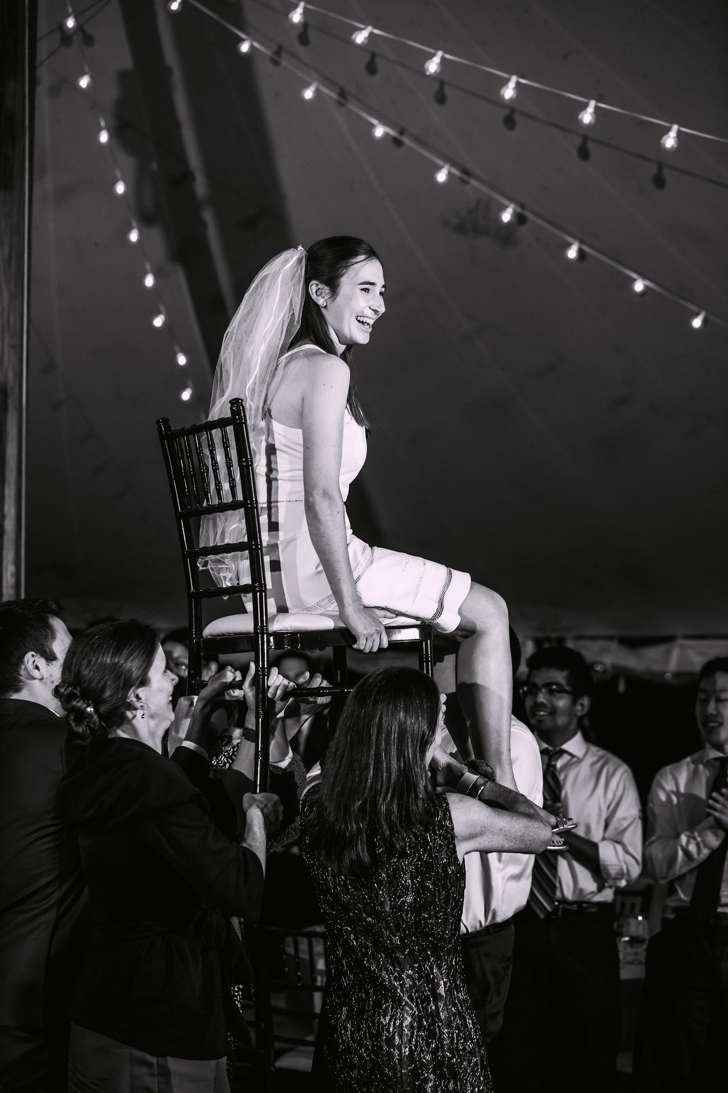 Black and white photo of a smiling bride lifted on a chair during a festive wedding hora under string lights, The Bradley Estate, Canton, MA