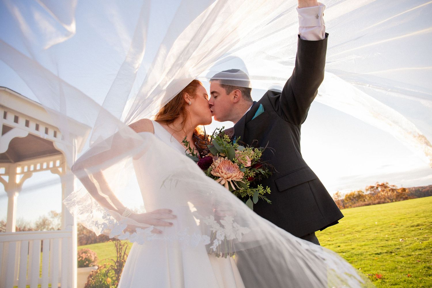 Bride and groom share a kiss under billowing veil near gazebo on wedding day, Xenia Helix Photography, Boston, MA