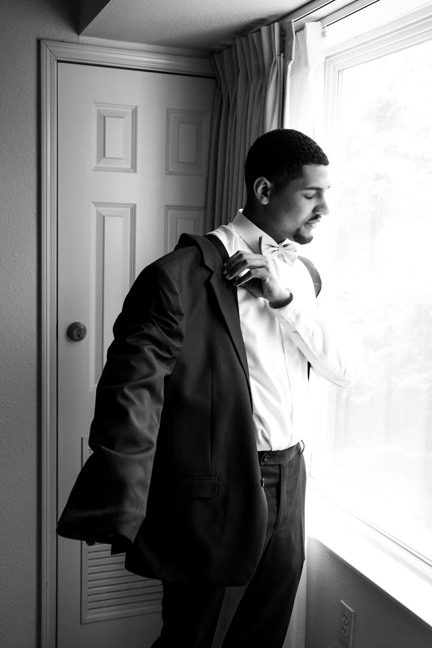 Dapper groom in tuxedo adjusting bow tie by window in elegant black and white photo, Groveland Fairways, Groveland, MA