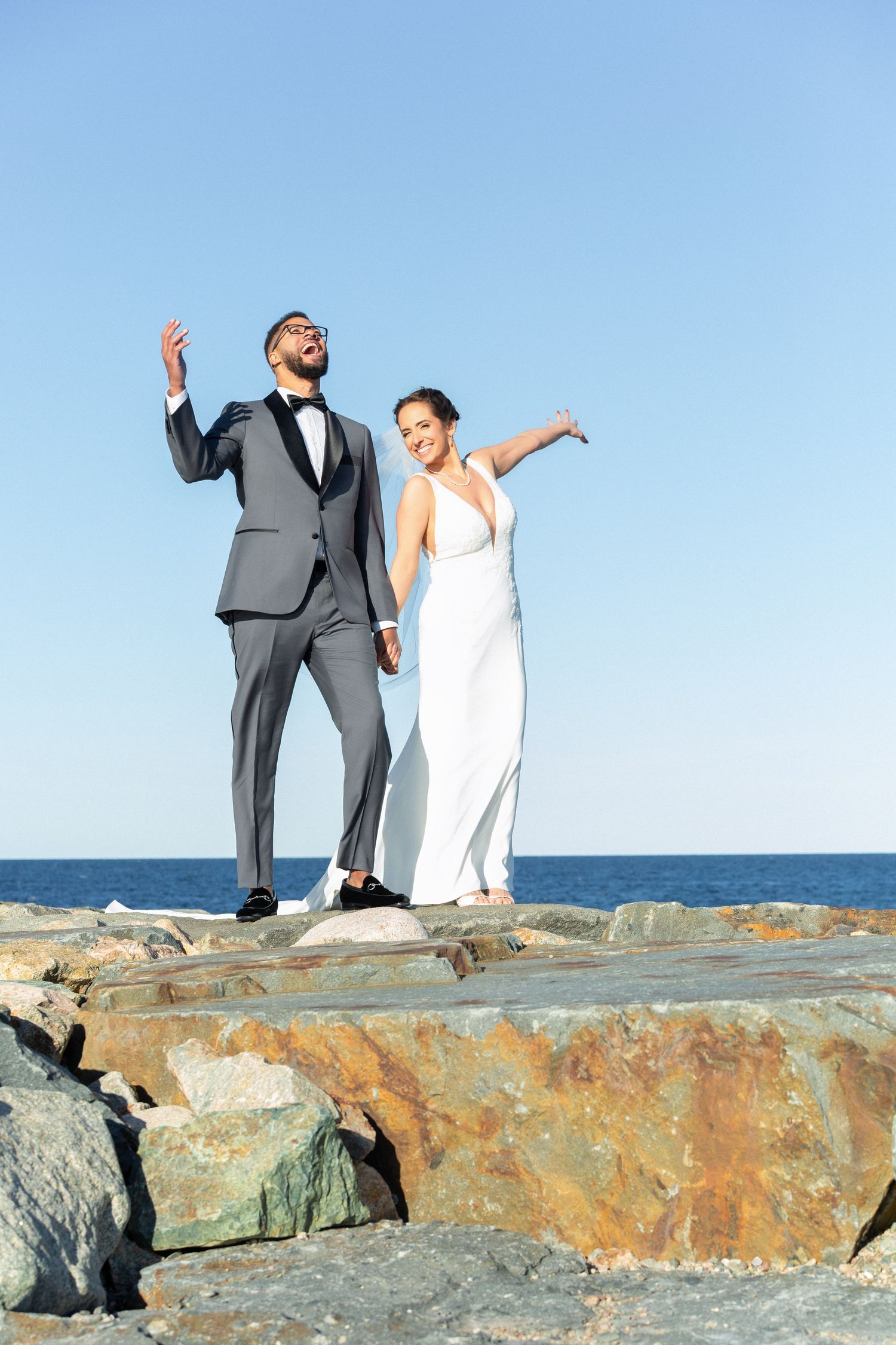 Joyful newlyweds stand on coastal rocks with arms raised celebrating their wedding by the sea, Scituate Lighthouse, Scituate, MA