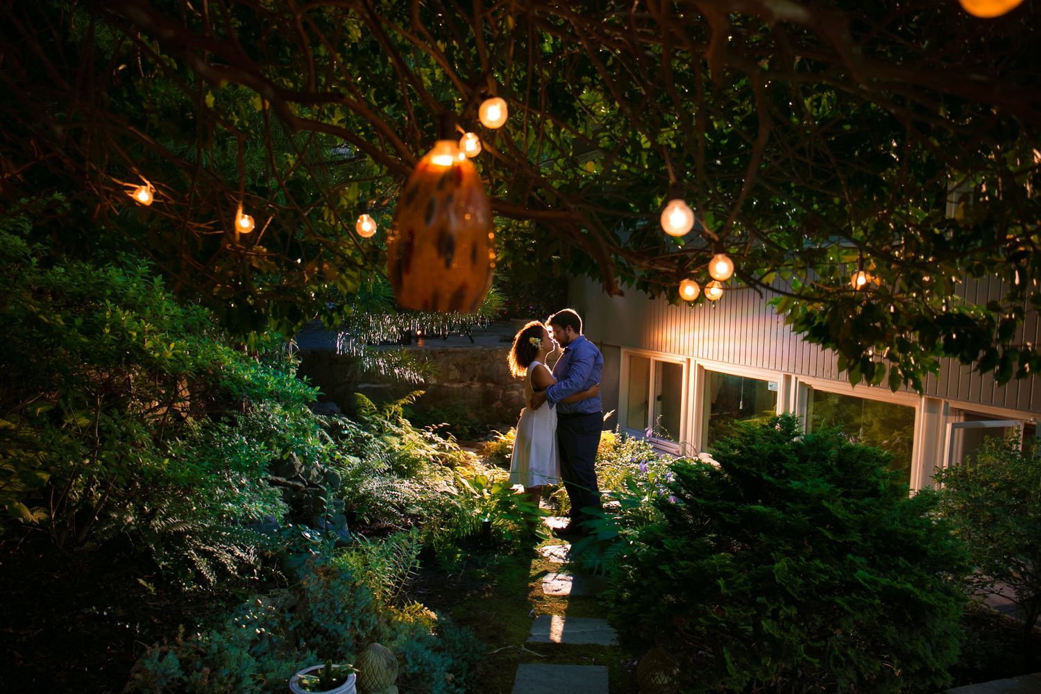 Couple sharing a romantic embrace under string lights in a lush garden during evening wedding reception, Xenia Helix Photography, Boston, MA