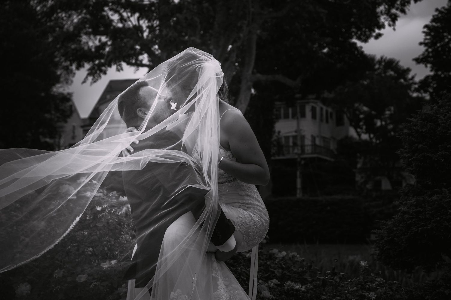 Black and white photo of bride and groom kissing under a flowing veil as he holds her in his arms, Brewster Gardens, Plymouth, MA