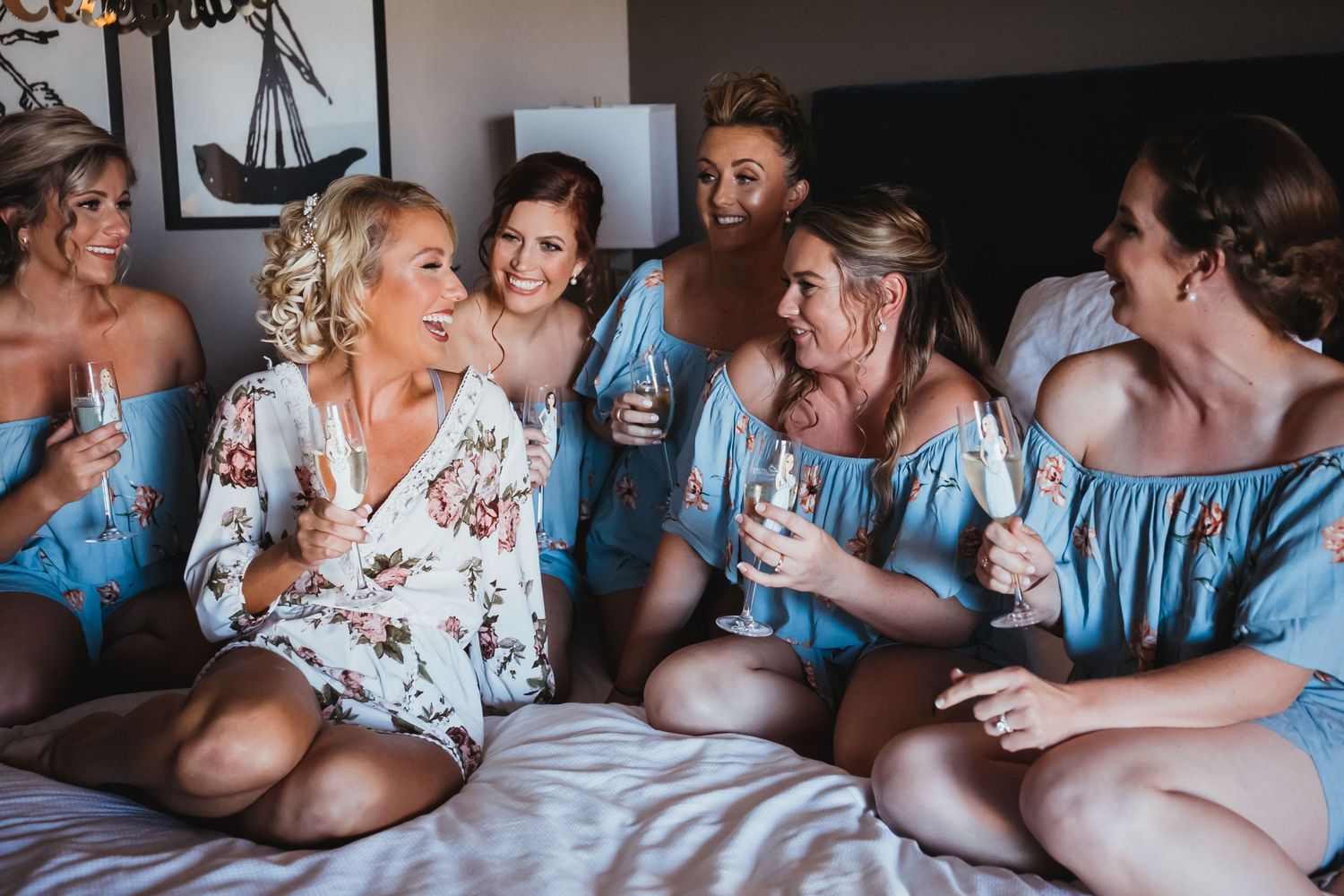 Bride in a floral robe celebrates with bridesmaids in blue dresses, holding champagne on bed, Hotel 1620 Plymouth Harbor, Plymouth, MA
