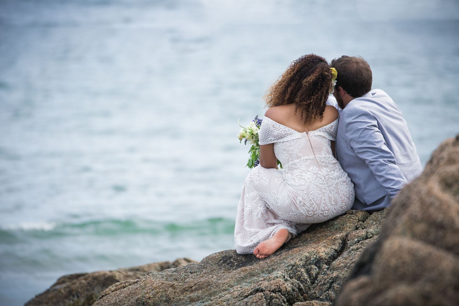 Newlywed couple sitting on coastal rocks overlooking the ocean, bride in white lace dress, Xenia Helix Photography, Boston, MA