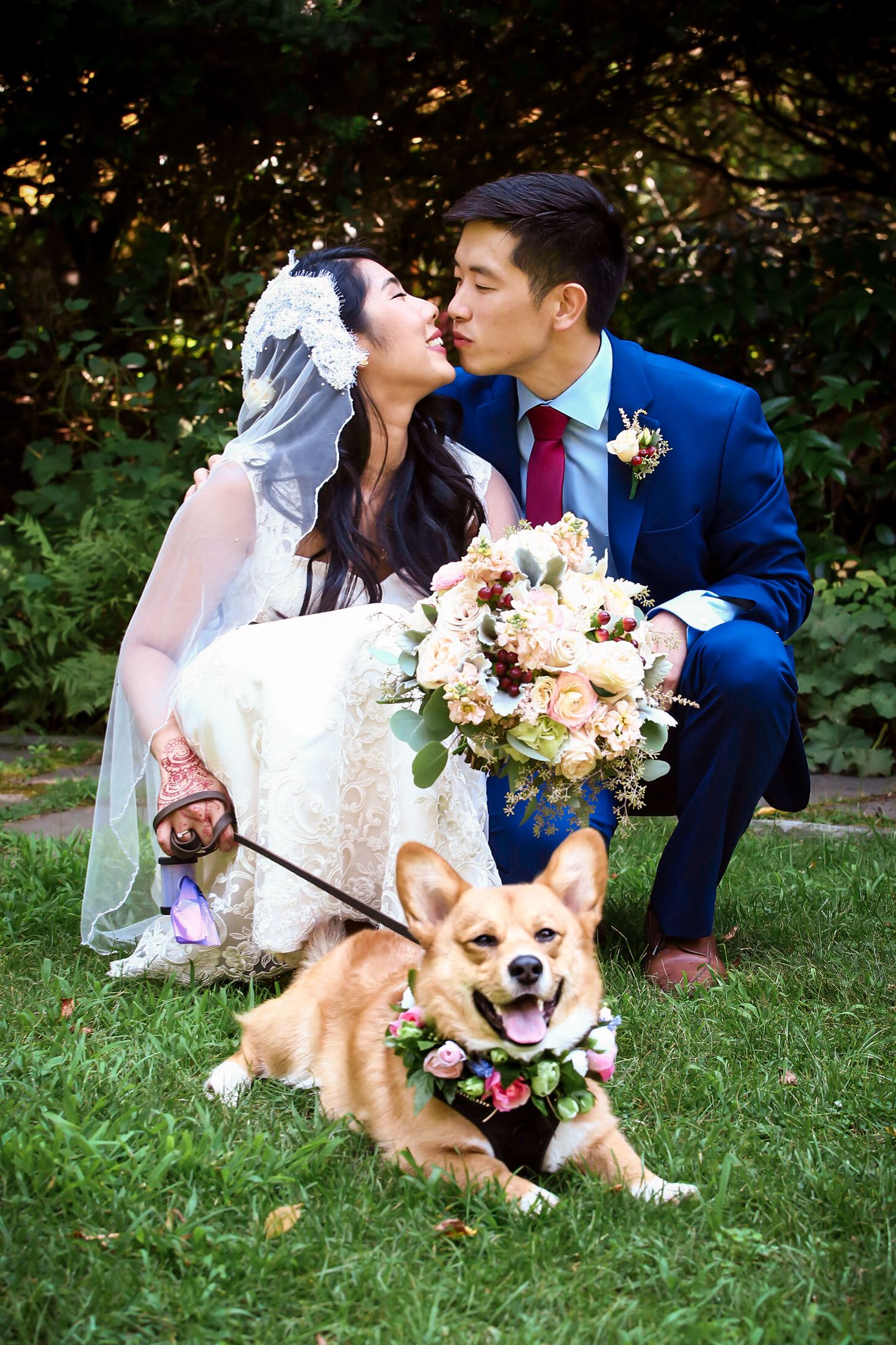 Bride and groom kiss while crouching beside their flower-crowned Corgi dog in garden setting, Mass Audubon's Habitat, Belmont, MA