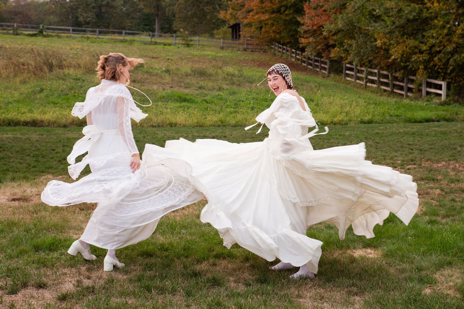 Two brides in flowing white wedding gowns twirl joyfully in an outdoor field with autumn foliage, Xenia Helix Photography, Boston, MA