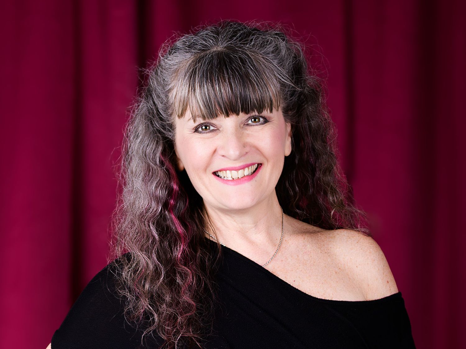 Smiling woman with curly dark hair and bangs wearing a black off-shoulder top against a red curtain backdrop.