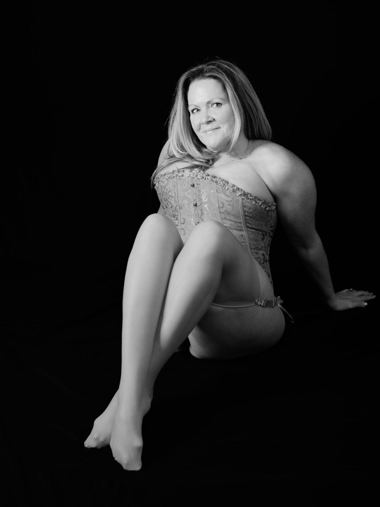 Black and white portrait of a smiling woman in a corset sitting gracefully against a dark background.