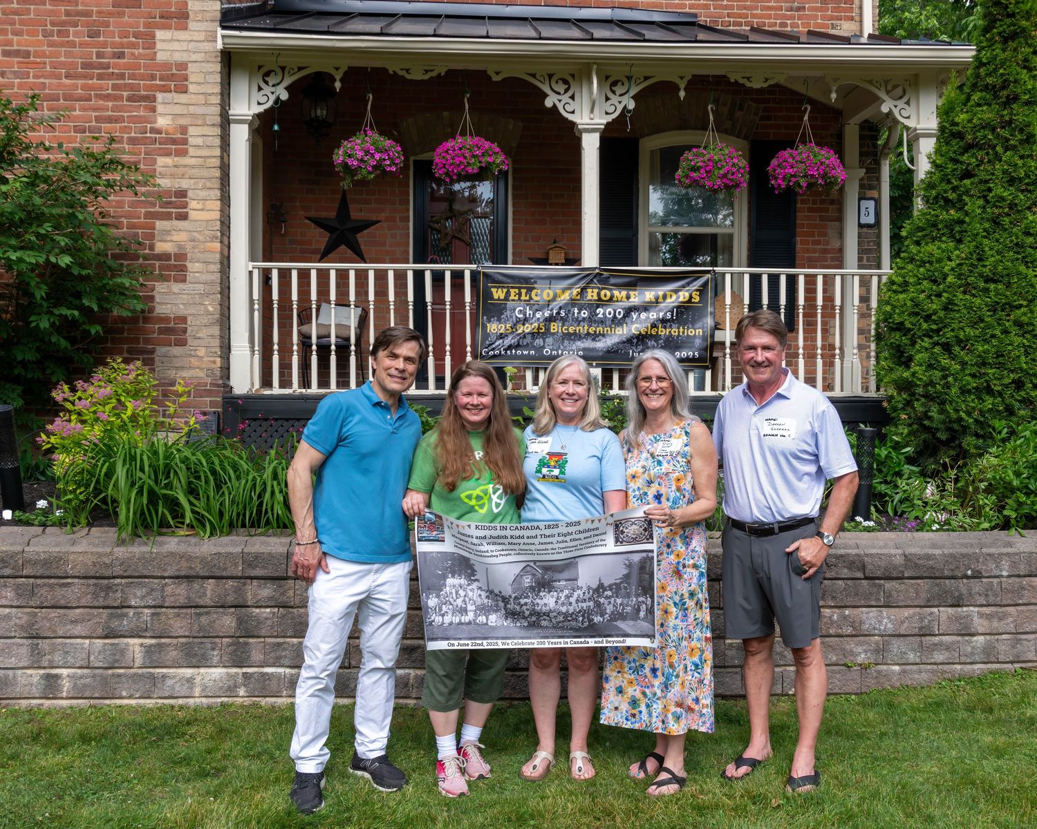 Five people pose outdoors holding a framed historic photo in front of a charming brick home with hanging flower baskets.
