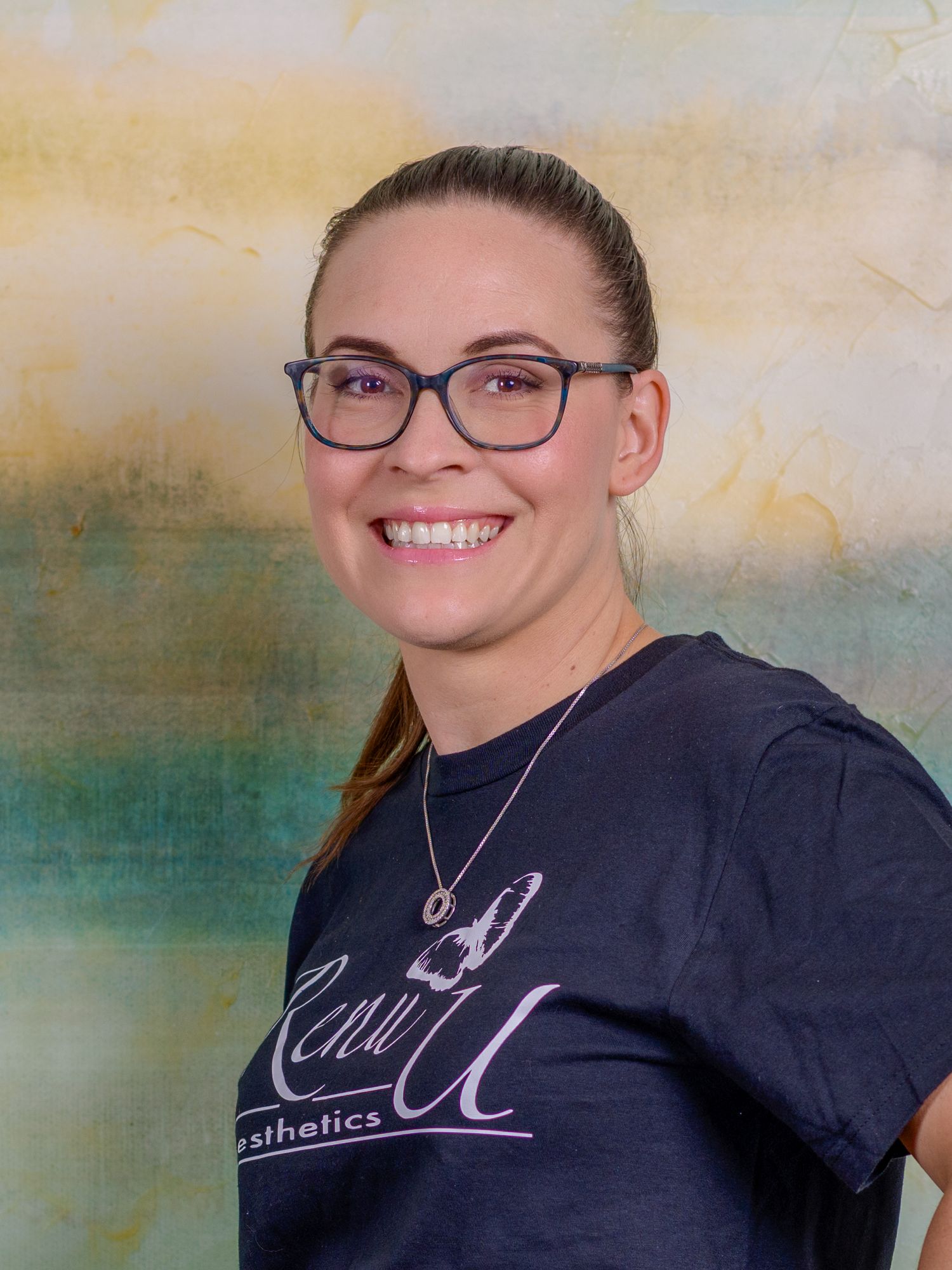 Smiling woman with glasses wearing a Renu U Aesthetics t-shirt poses for a professional headshot photo.