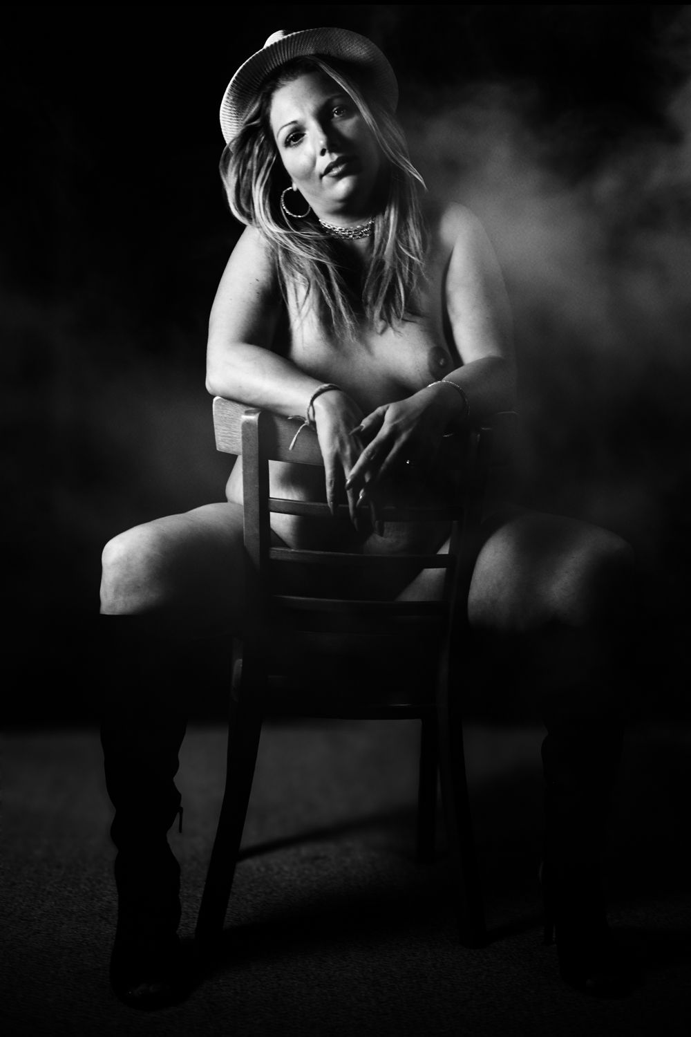 Black and white boudoir photo of woman in cowboy hat seated on chair with dramatic smoky background.