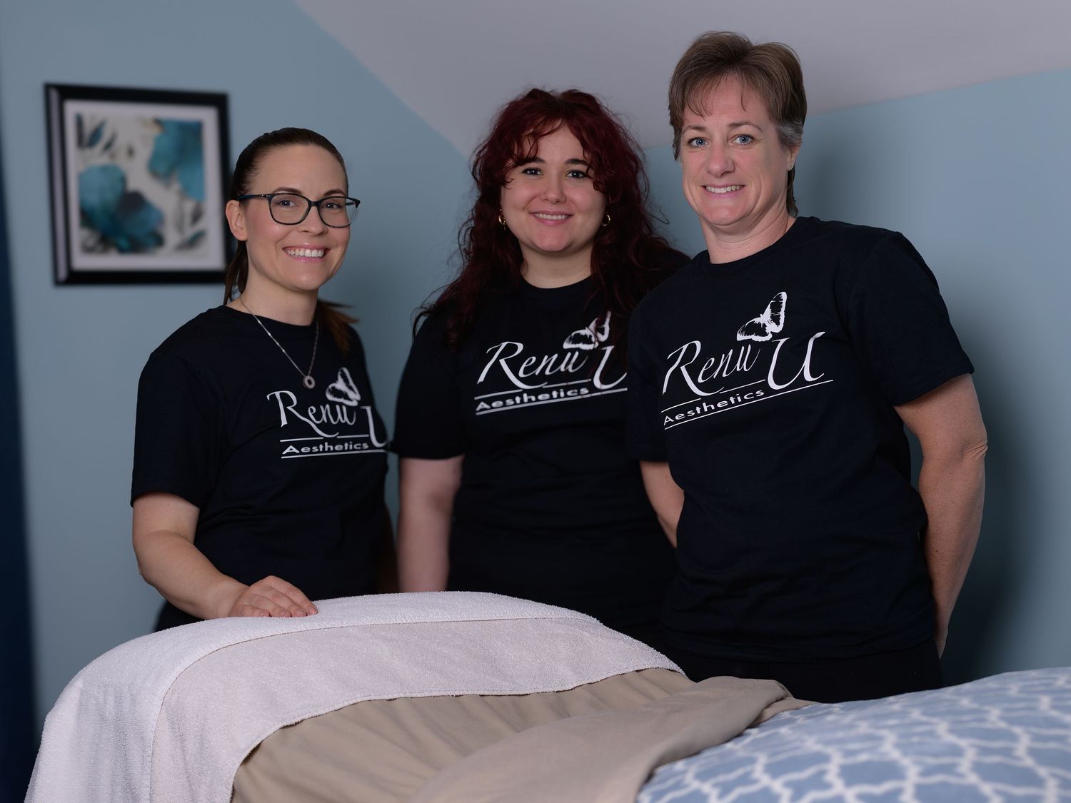 Three women in matching Renu U Aethetics black t-shirts smile together in a spa treatment room.
