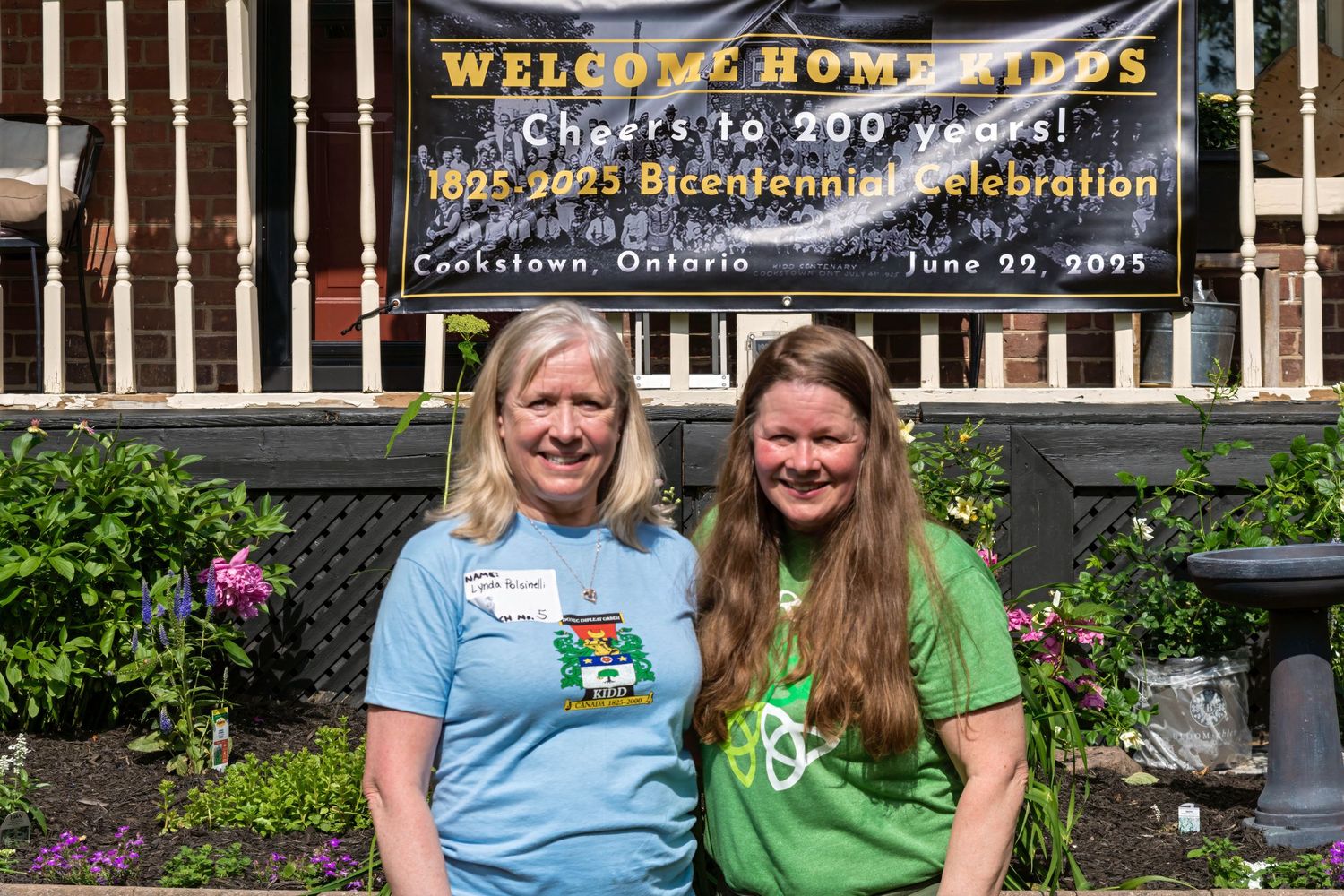 Two women smiling in front of a Welcome Home Kids Bicentennial Celebration banner in Cookstown, Ontario.
