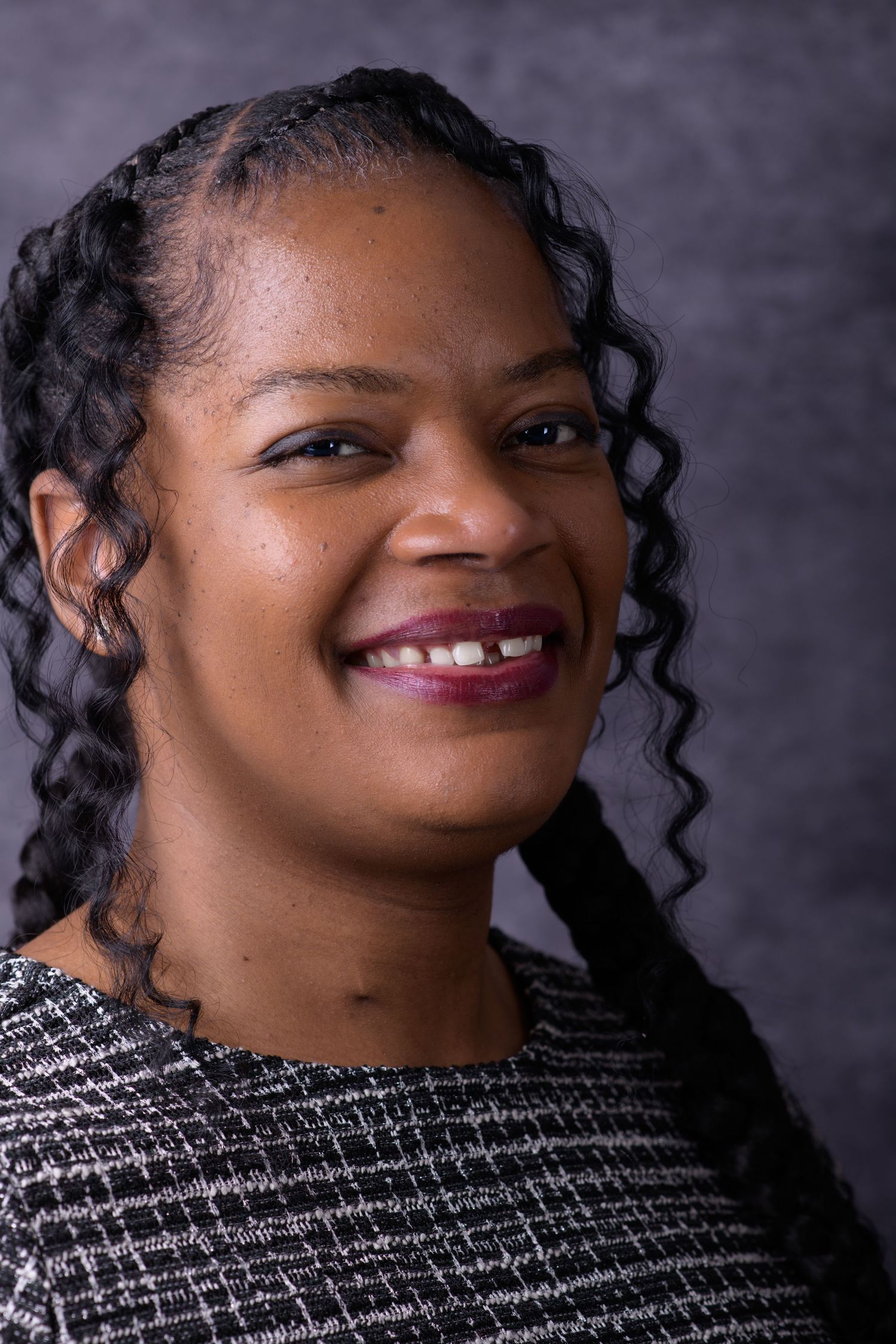 Smiling Black woman with curly hair wearing a patterned blazer, posing for a professional portrait against a gray background.