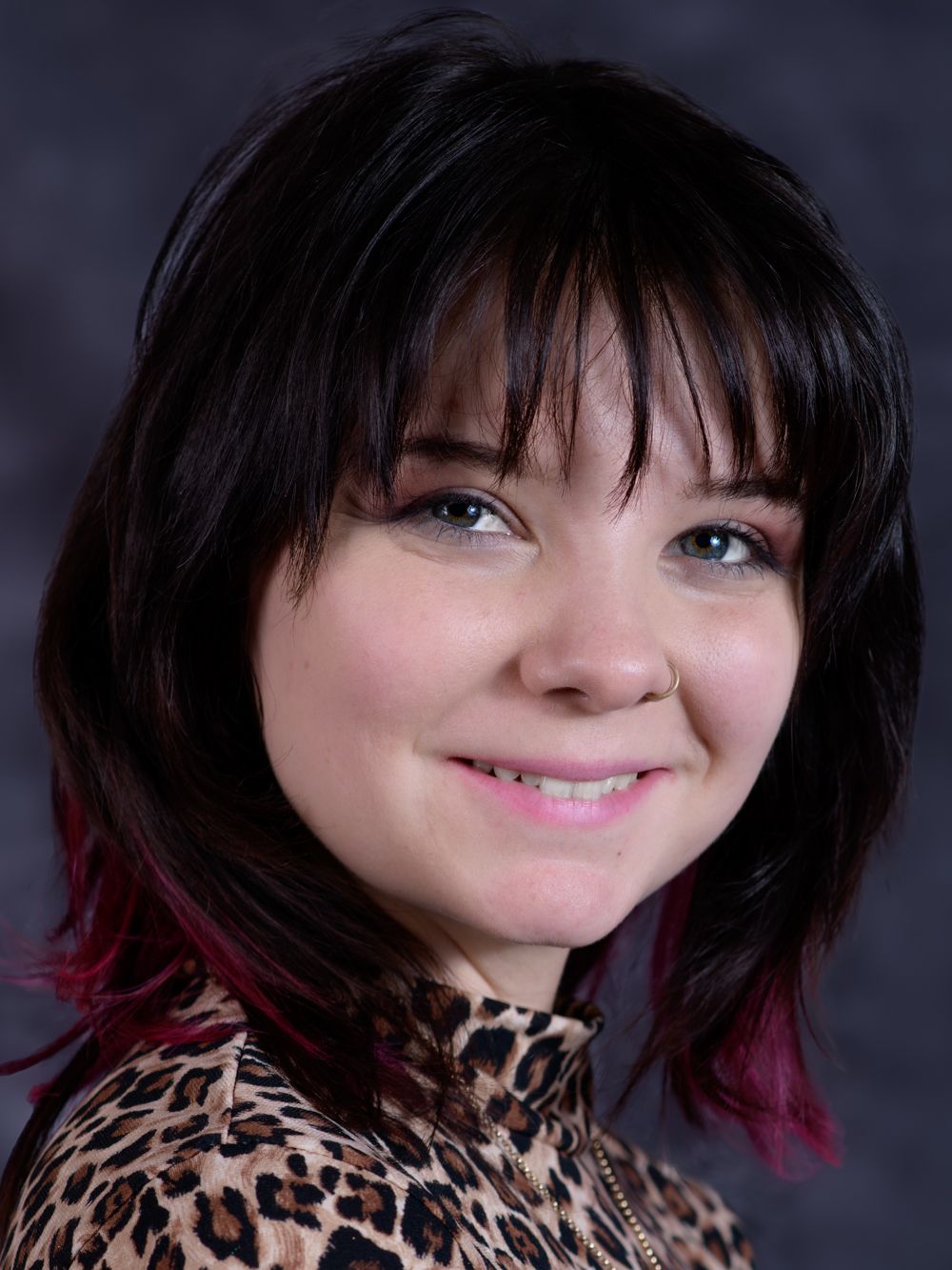 Young woman with dark hair and pink highlights wearing leopard print top in a professional headshot.