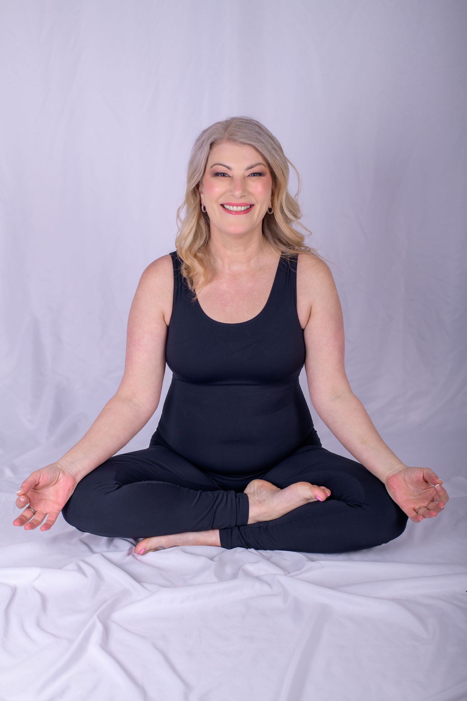 Smiling mature woman in black yoga outfit sitting in lotus meditation pose on white fabric background.