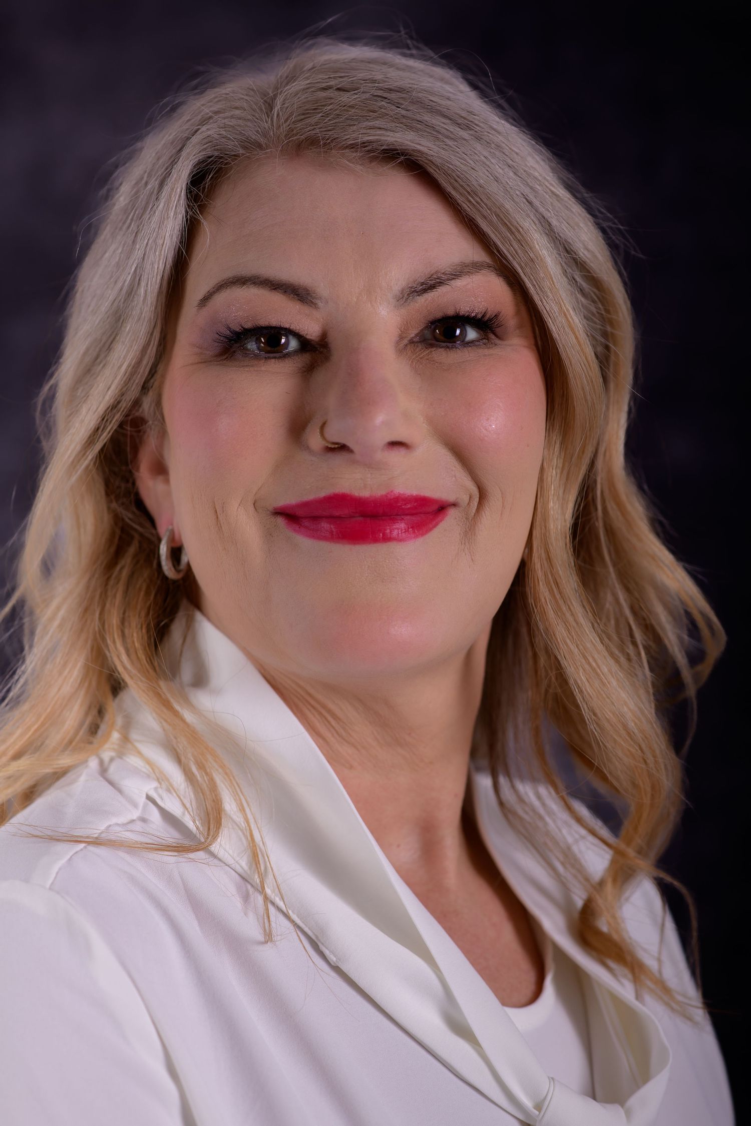 Professional headshot of a smiling blonde woman in a white blazer with red lipstick against a dark background.