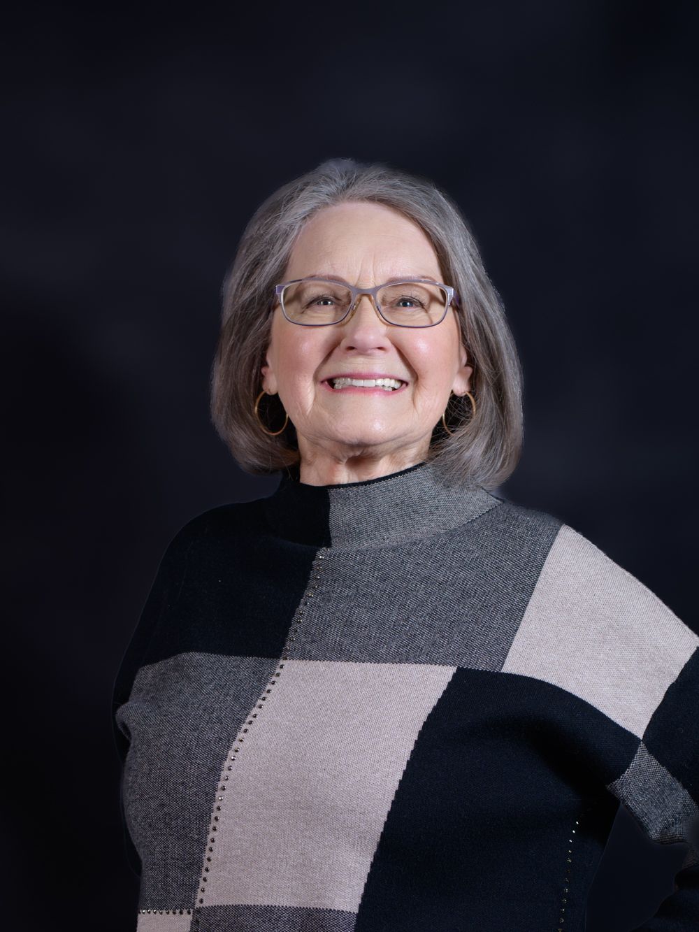 Smiling older woman with gray hair and glasses wearing a black and gray color-block turtleneck sweater.