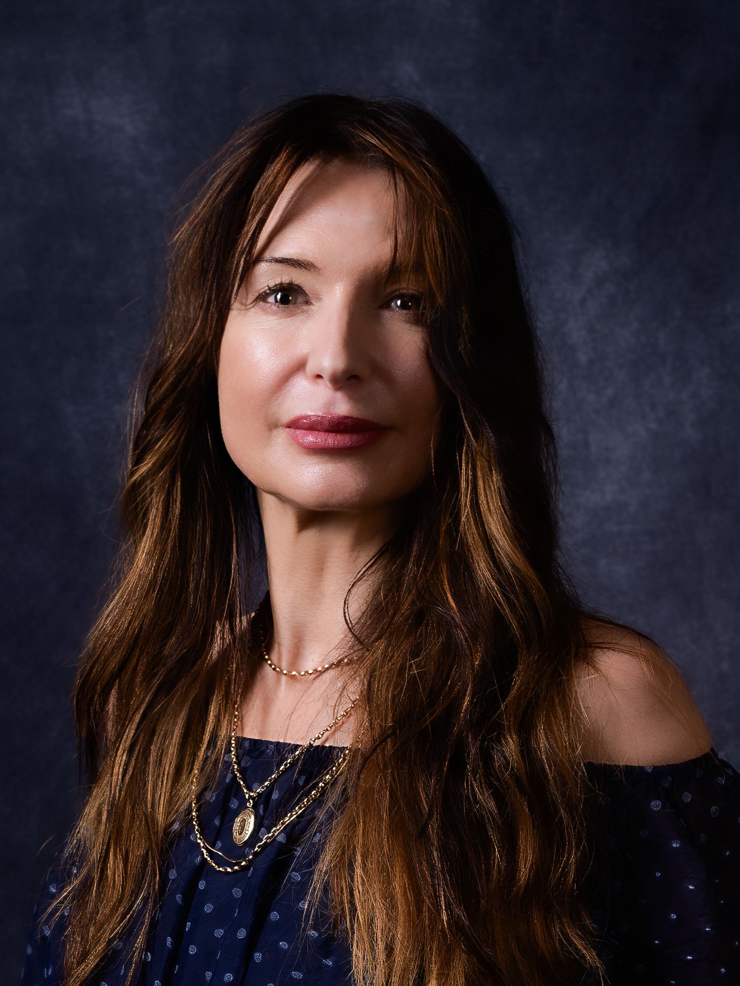 A woman with long brown hair wearing a navy off-shoulder top and gold necklaces poses for a professional portrait.