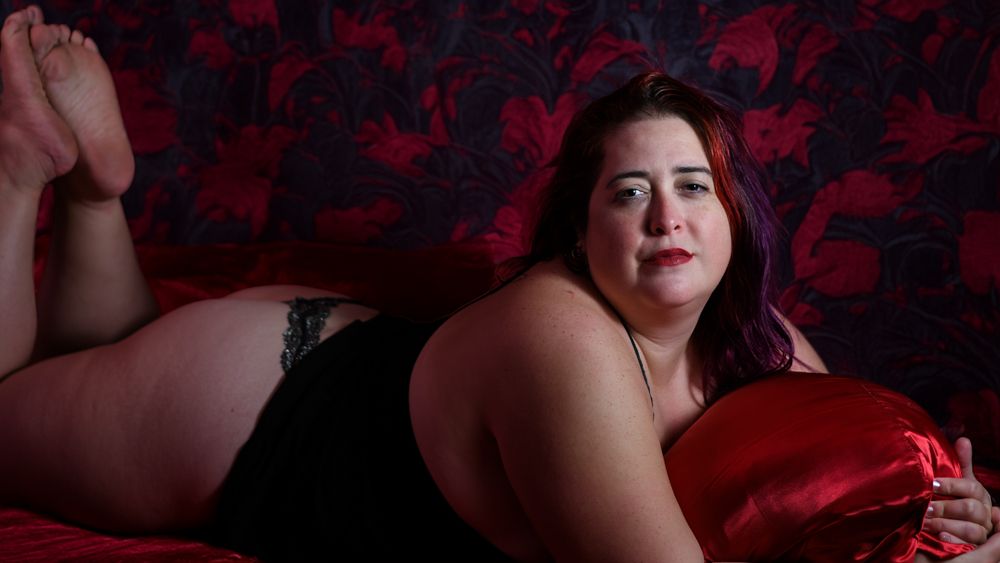 A plus-size woman in black lingerie poses on a red bed against a dark floral wallpaper backdrop.