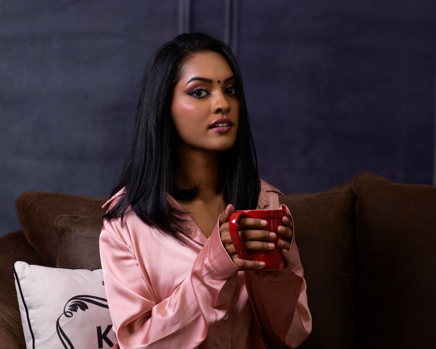 Young woman in pink satin robe holding red mug, sitting on brown sofa with dramatic eye makeup.
