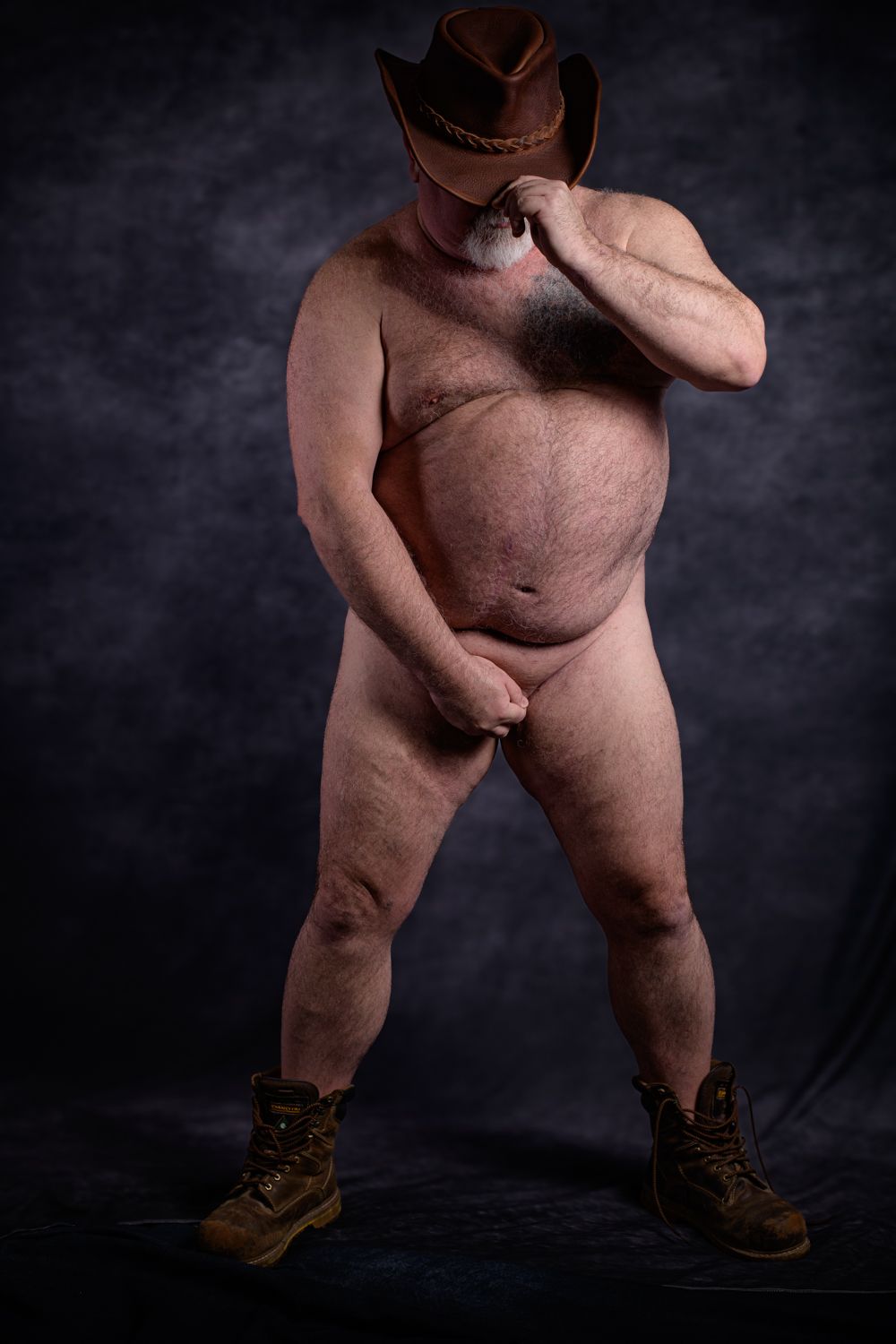 A nude man at a studio shoot wearing a brown cowboy hat, looking down with hands positioned for modest coverage.