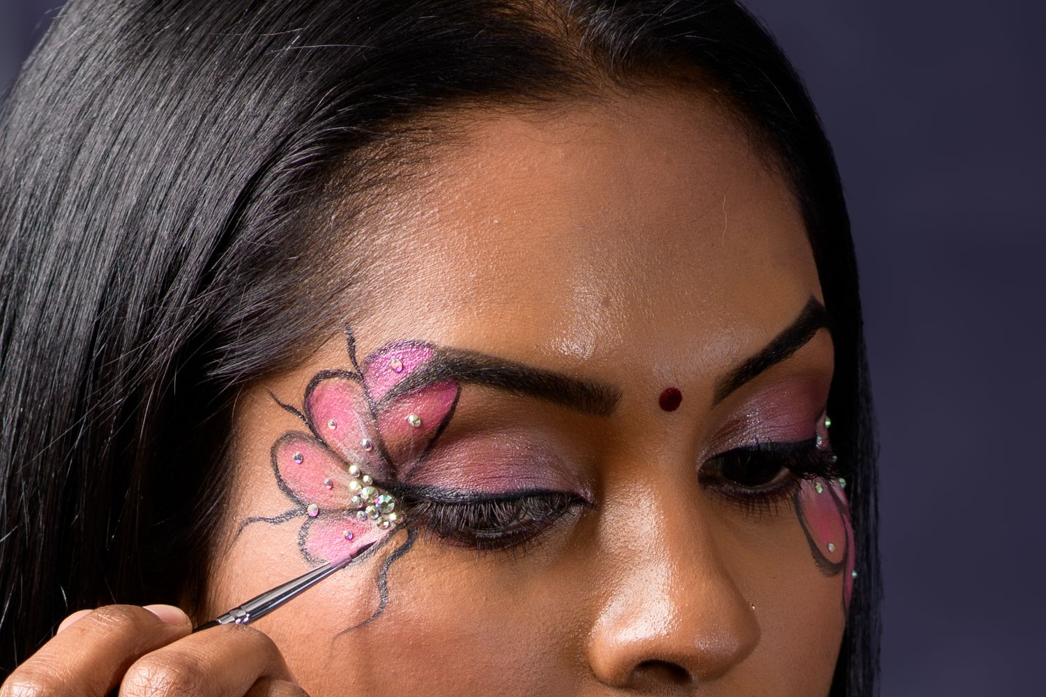 Artist applying intricate pink butterfly eye makeup with a brush on a woman with dark hair.