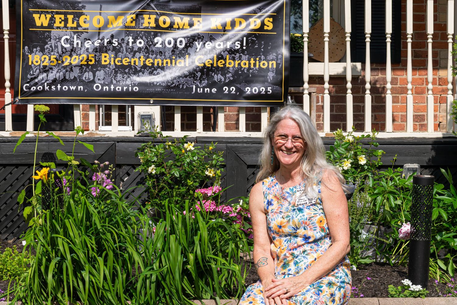 Woman sitting in front of a Welcome Home banner for Cookstown Ontario Bicentennial Celebration 1825-2025.
