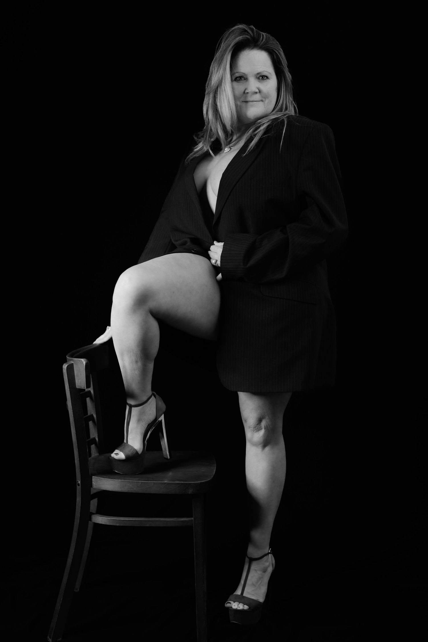 Black and white studio portrait of a woman in a blazer posing on a chair with high heels.