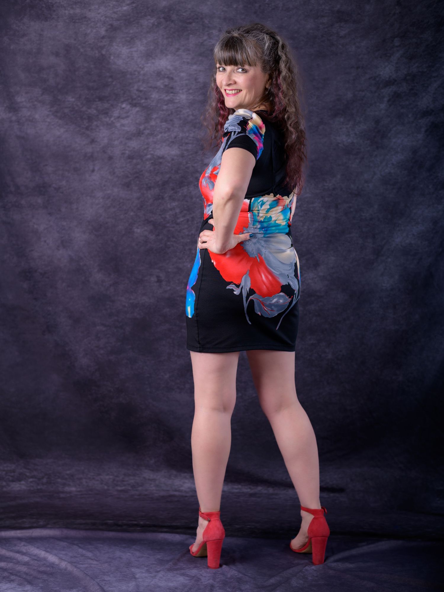 Woman in floral black mini dress and red heels poses confidently against dark studio backdrop.
