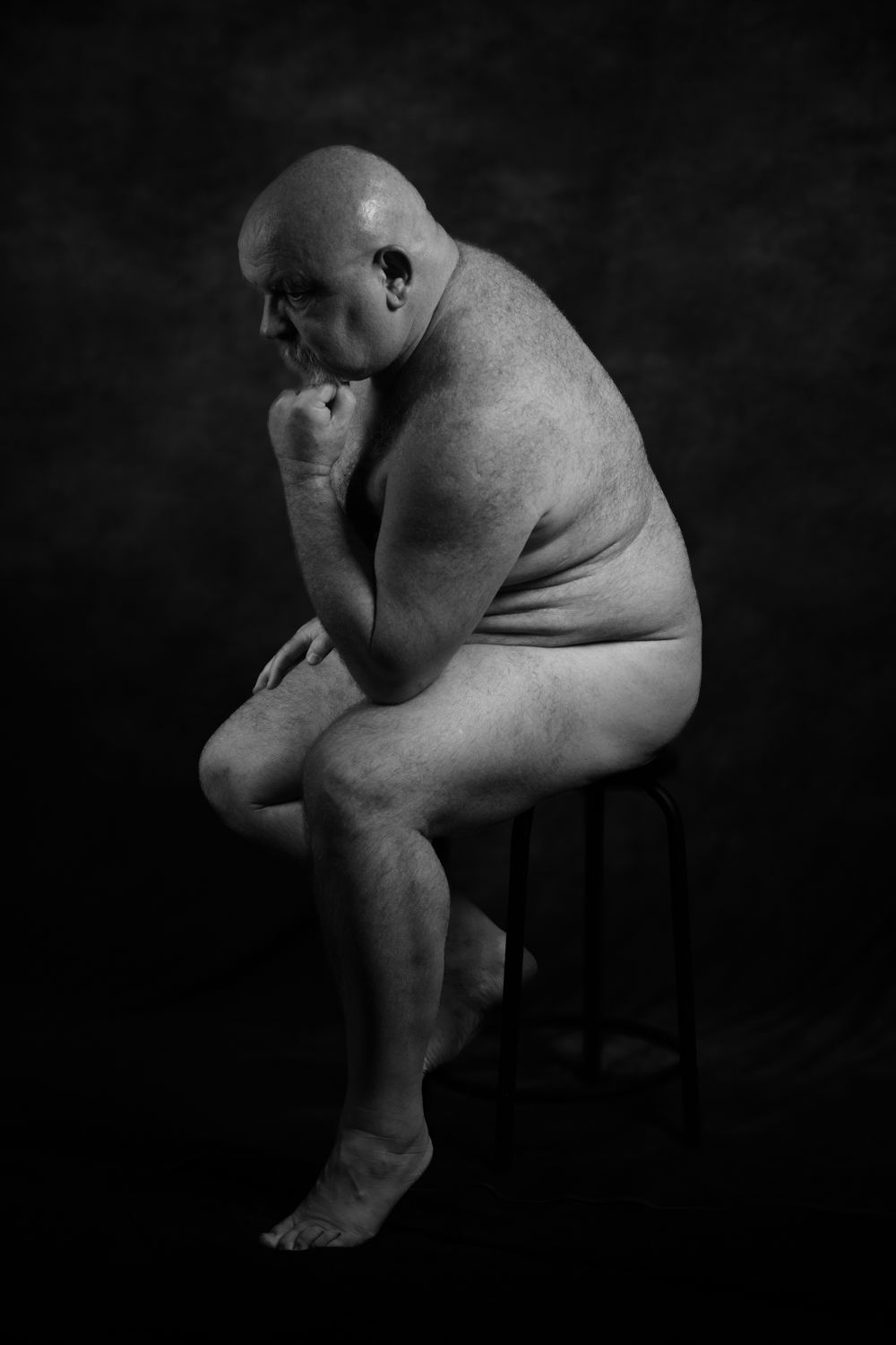 A deep black and white side profile of a man sitting on a stool in a contemplative pose similar to Rodin’s Thinker.