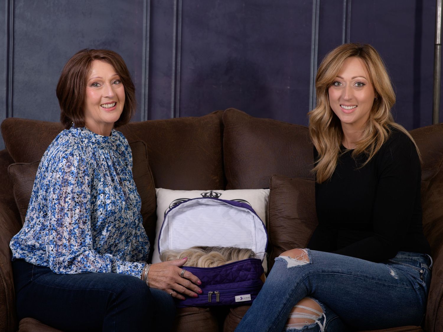 Two women sitting on a brown leather sofa smiling with a decorative pillow featuring a purse design between them.