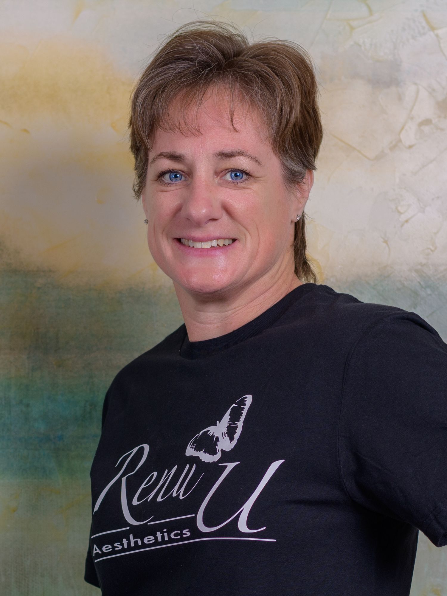 Woman in black Renu U Aesthetics t-shirt smiling confidently against a soft green and beige background.