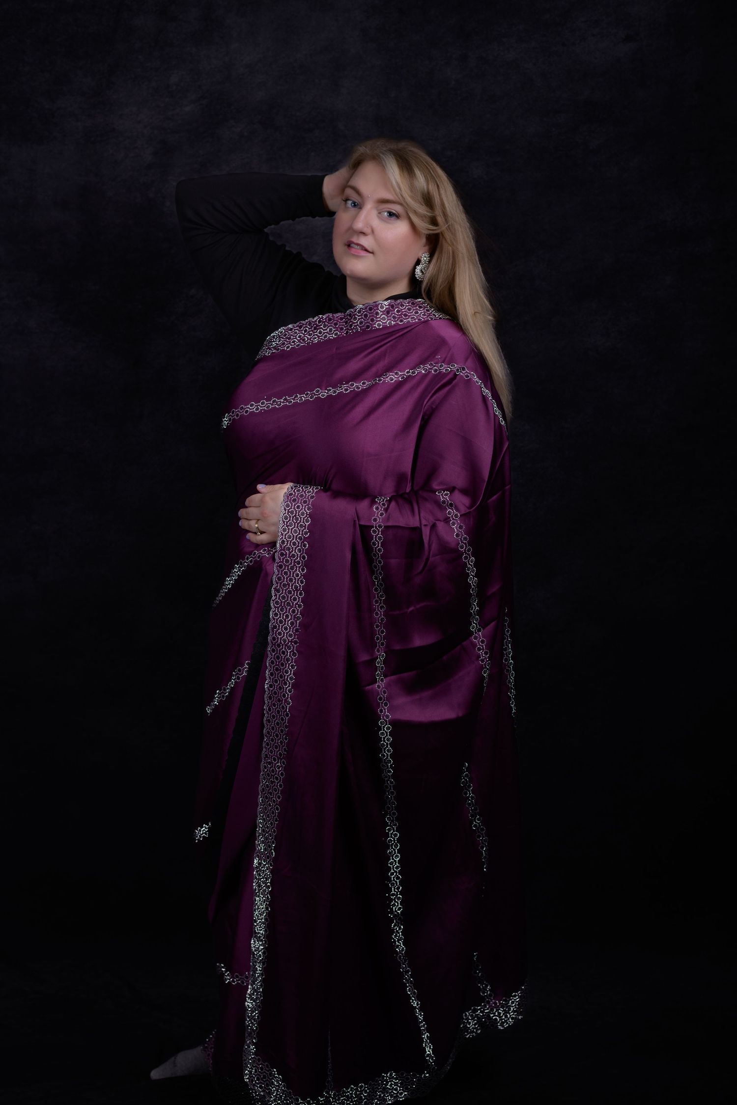Woman in elegant purple sari with silver trim poses gracefully against dark studio background.