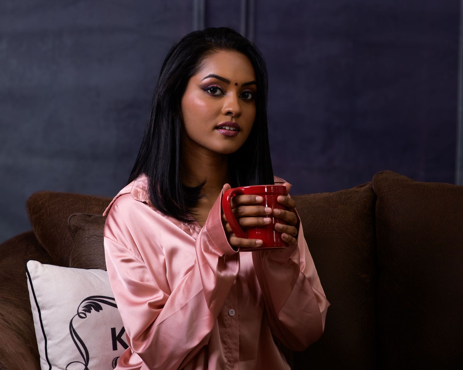 Young South Asian woman in pink satin pajamas holding a red mug while sitting on a brown couch.