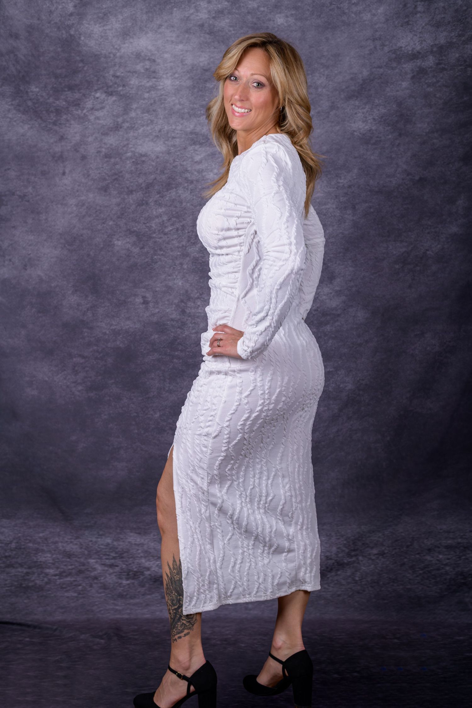 Smiling blonde woman posing in a white long-sleeve textured midi dress with a slit and black heels.