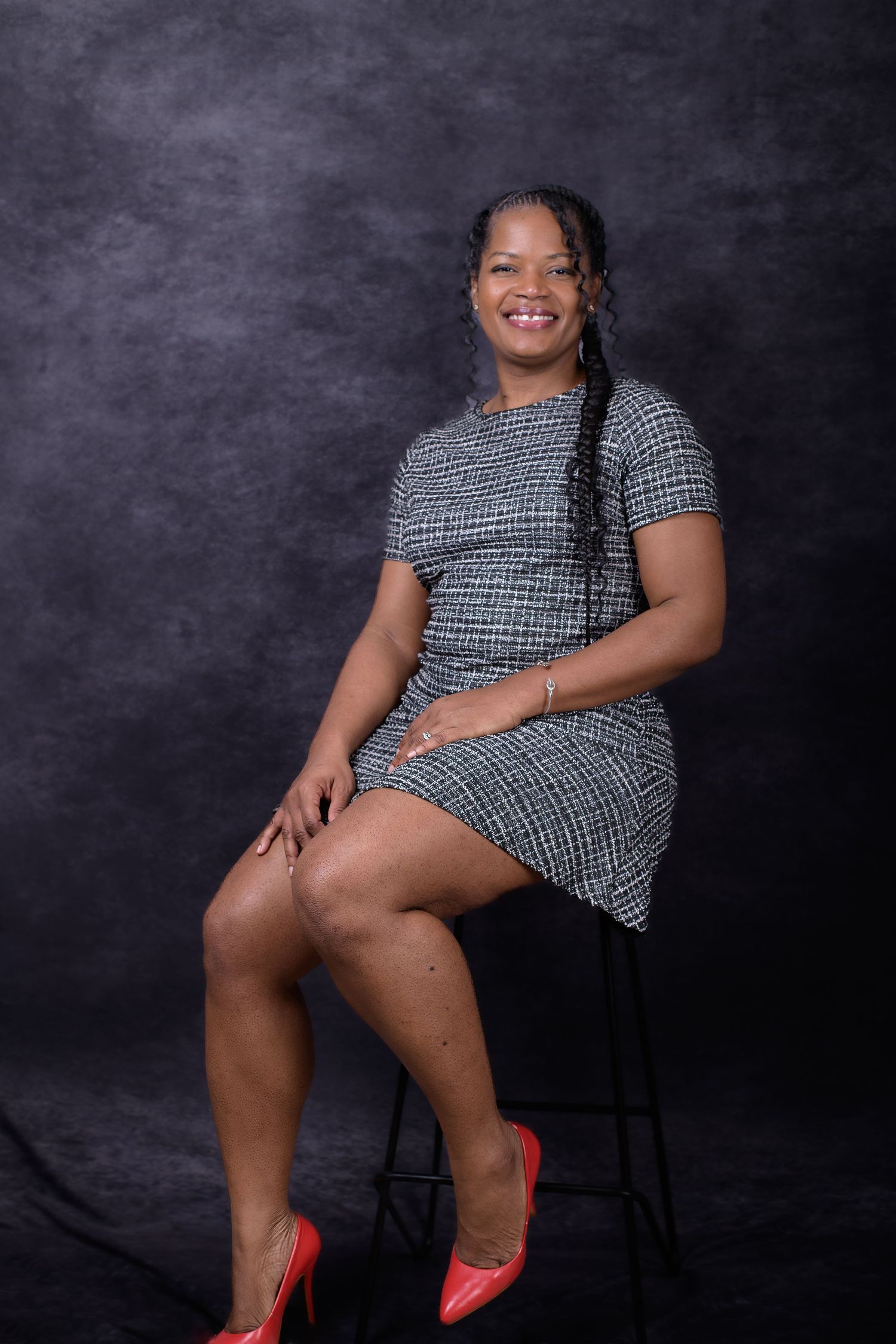 Woman smiling in gray tweed dress seated on stool with red heels against dark studio background.