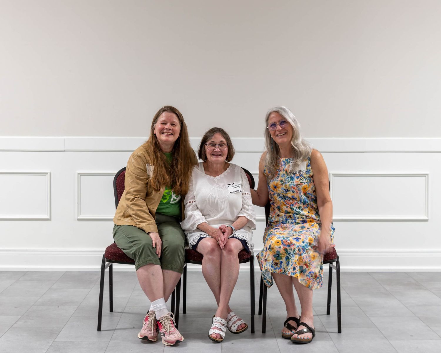 Three women sitting on chairs smiling together in a bright white room.