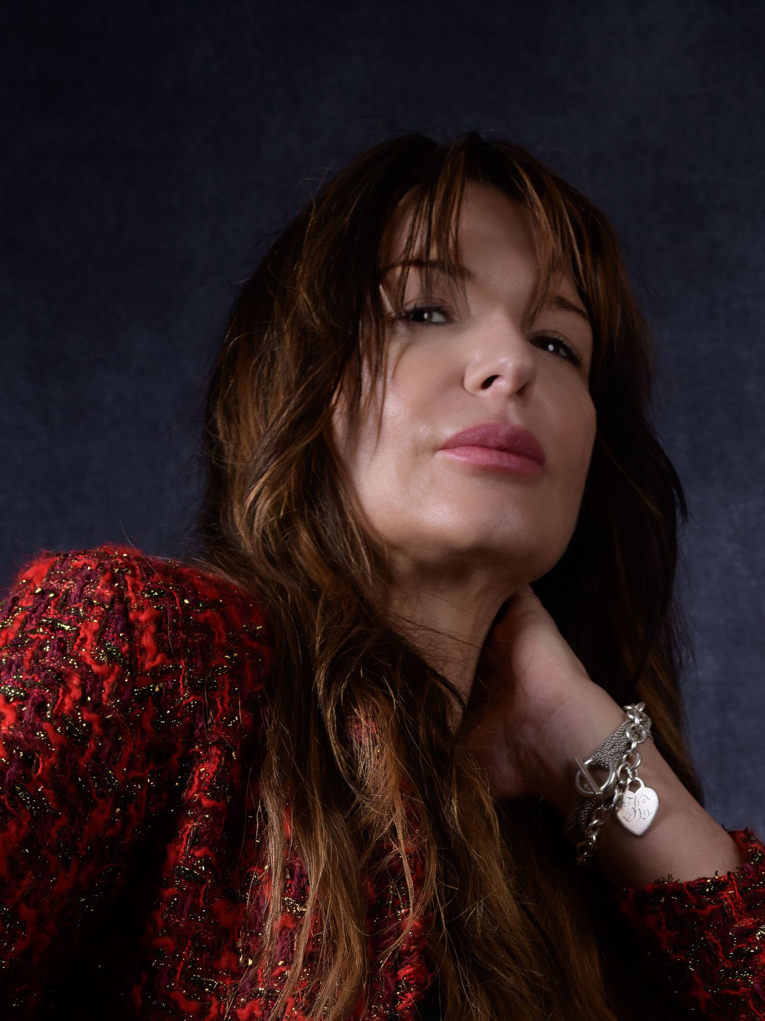 Woman with long brunette hair in red tweed jacket poses elegantly, touching her neck, wearing a charm bracelet.