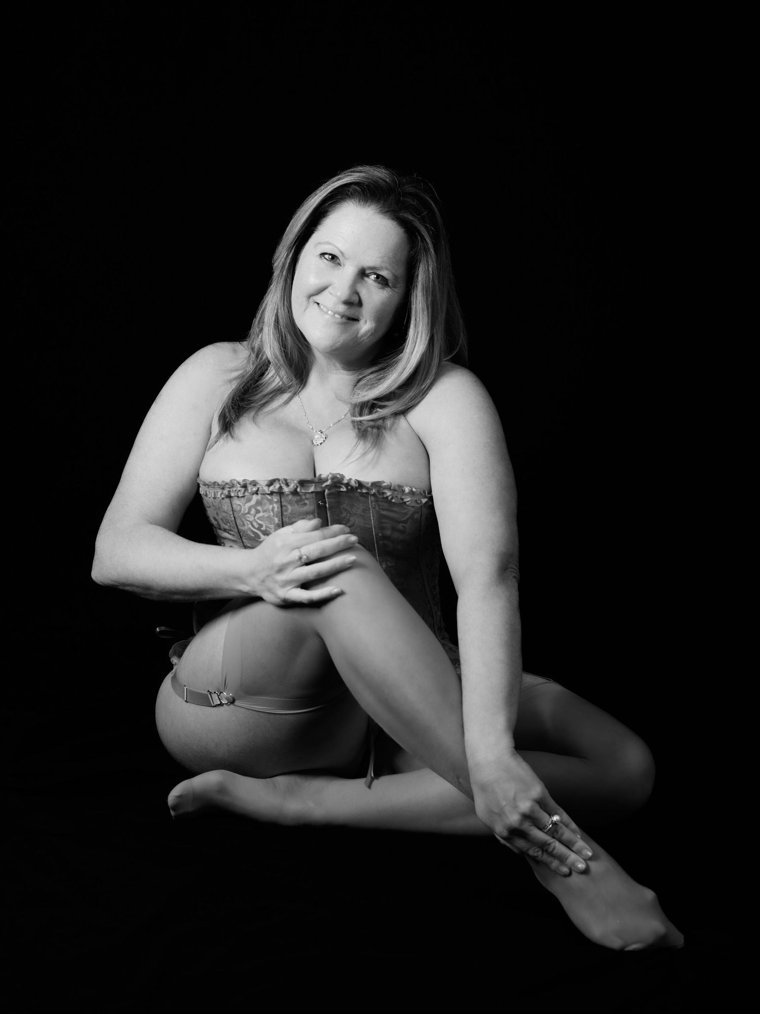 Black and white boudoir portrait of a smiling woman in a corset, seated gracefully against a dark studio background.