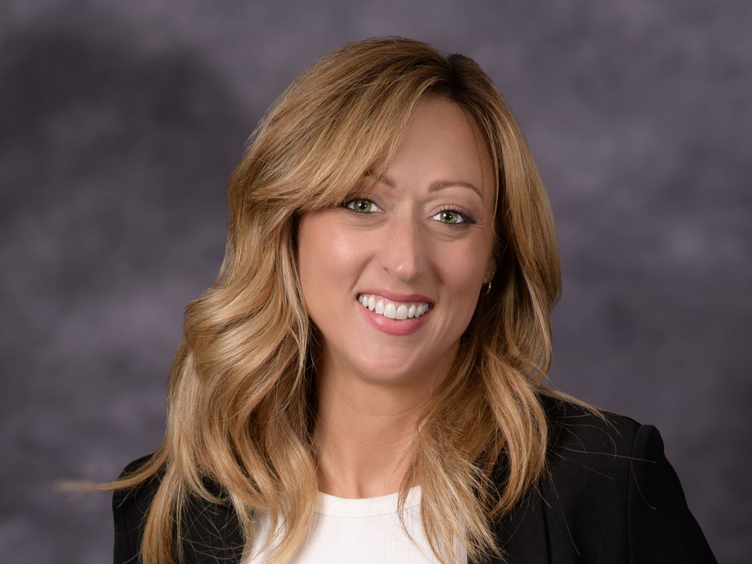 Professional headshot of a smiling woman with blonde wavy hair wearing a black blazer against a gray background.
