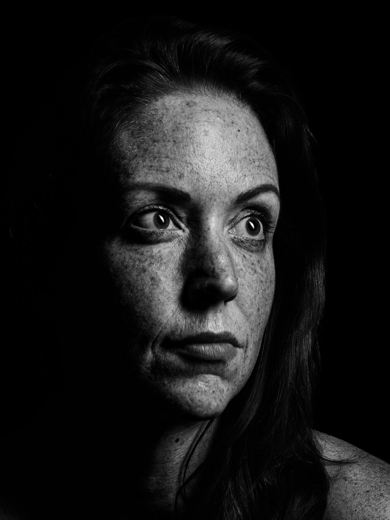 Black and white portrait of a freckled woman with intense eyes against a dark background.