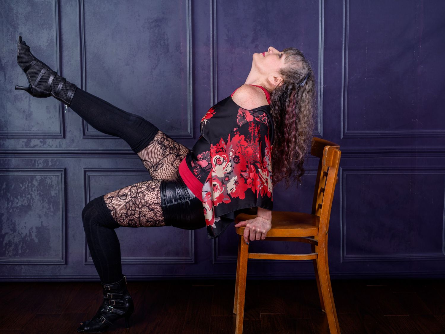 Woman in red floral top and black lace tights poses dramatically on wooden chair with leg raised high.